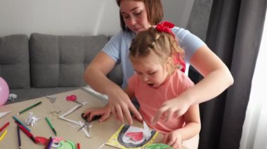 Annenin orta pozu, bir çocuğun tatil kartı oymayı öğrenmesine yardımcı olur. Küçük beyaz bir kız Anneler Günü ve Çocuklar Günü için el işi yapıyor..