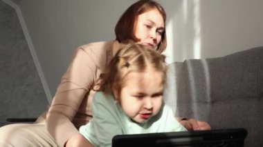 Yetişkin beyaz bir anne, saçlarını şirin küçük kızının saç stiline örer. Çocuk bir tablet kullanıyor ve bir çizgi film izliyor. Birlikte geçirdiğimiz aile zamanı..