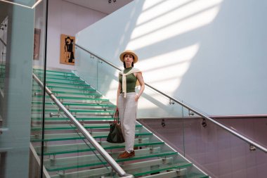 Hasır şapkalı, şeffaf cam merdivenlerden inen bir çanta taşıyan beyaz, zarif bir kadının geniş görüşü. Duvarda güneş ışınları.