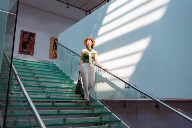 Hasır şapkalı, şeffaf cam merdivenlerden inen bir çanta taşıyan beyaz, zarif bir kadının geniş görüşü. Güneş ışınları duvarda. İş konsepti.