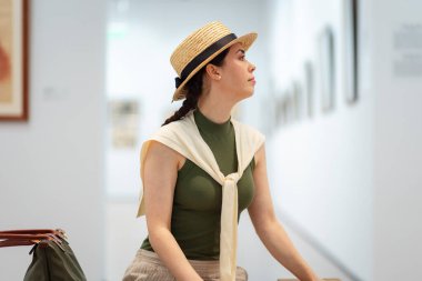 Hasır şapkalı ve zarif elbiseli güzel beyaz bir kadının çağdaş bir sanat galerisini ziyaret edişinin orta fotoğrafı. Yan görüş. Müze sergisi.