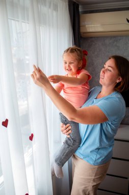 Beyaz bir anne küçük kızını tutuyor ve perdeleri kırmızı kağıt kalplerle süslüyor. Dikey. Sevgililer Günü ve Anneler Günü için kutlama.