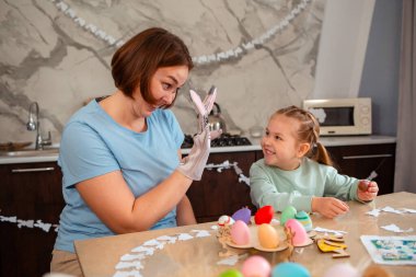 Mutlu anne ve küçük gülümseyen kızı Paskalya bahar tatili için çikolata yumurtalarını açıp oynuyorlar. Elinde tavşan eldiveni olan beyaz bir kadın bir çocukla oynuyor..