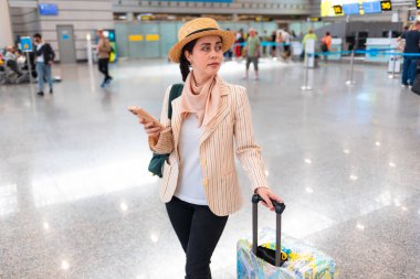 Hasır şapkalı, akıllı telefon kullanan ve bavulunu havaalanında ya da istasyonda taşıyan beyaz, zarif bir kadının portresi. Online kontrol uçuşu. Tatil ve gezi konsepti.