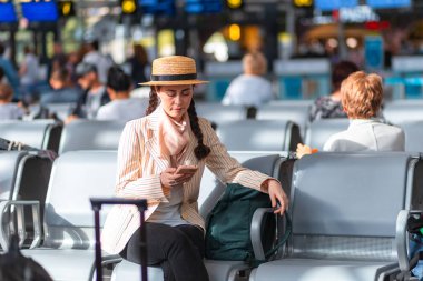Zarif giyimli genç beyaz kadın havaalanında ya da tren istasyonunda sandalyelerde oturuyor. Akıllı telefon kullanan bir kadın. Seyahat ve tatil kavramı.