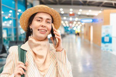 Şapkalı ve ceketli genç beyaz kadın portresi havaalanındaki akıllı telefona gülümsüyor ve onunla konuşuyor. Uzayı kopyala Kameraya bakan kişi. Uçuştan önce terminalde biniş kavramı.