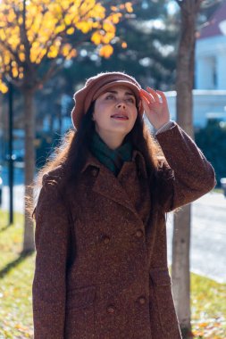 Güneşli bir şehirde sonbahar caddesinde şapkası ve paltosuyla yürüyen güzel, genç, mutlu, beyaz bir kadının resmi. Dikey oryantasyon. Psikoloji kavramı ve pozitif ruh hali.