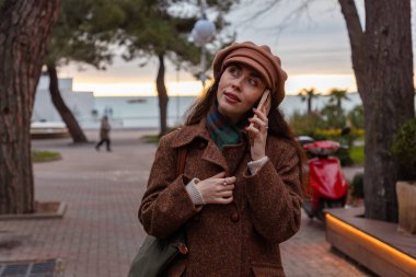 Şapkalı ve paltolu güzel beyaz bir kadının portresi sokakta yürüyor, gülümsüyor ve telefonda konuşuyor. Modern iletişim kavramı.