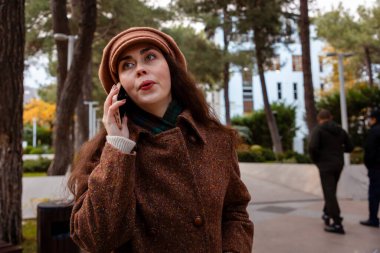 Şapkalı ve paltolu güzel beyaz bir kadının görüntüsü inanılmaz ve telefonda konuşuyor. Modern iletişim ve iş.