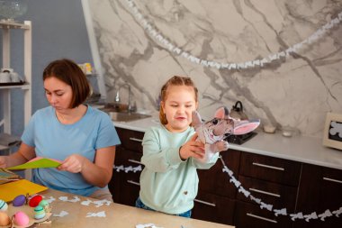 Genç anne ve mutlu küçük kızın Paskalya masasında oturan tavşan eldiveniyle oynadığı bir kare. Bahar tatili.