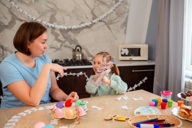 Gülümseyen anne ve tatlı küçük kız tavşan eldiveniyle oynuyorlar, Paskalya masasında oturuyorlar. Bahar tatili.