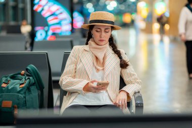 Beyaz tenli zarif bir kadının orta pozu havaalanı şezlongunda oturuyor ve akıllı telefon kullanıyor. Uçuş uygulaması kavramı.