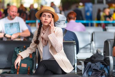 Şapka ve ceket giymiş genç beyaz bir kadın bekleme odasında bir koltukta oturuyor ve akıllı bir telefonla konuşuyor. Uçağa binmek için bekliyor..