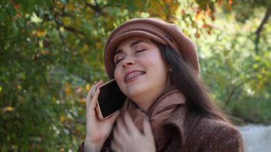 Şapkalı ve paltolu mutlu beyaz bir kadının portresi telefonda parkın arka planında konuşuyor. Kötü haber, şok ve heyecan ifadeleri.