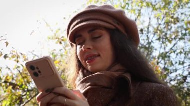 Parkta akıllı telefon kullanan gülümseyen genç bir kadın manzarası. Çevrimiçi sosyal ağlarda neşeli iletişim kavramı.