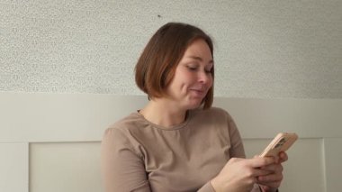 Genç gülümseyen bir kadının portresi evde koltukta otururken bir akıllı telefon kullanır. Sosyal medya ve çevrimiçi uygulamalar kavramı.