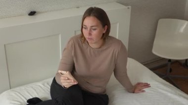 Kafkasyalı genç bir kadının en iyi görüntüsü evinde yatağında akıllı telefonunu kullanıyor. Çevrimiçi uygulamalar ve serbest çalışma kavramı.