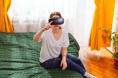 Kafkasyalı bir kadın yatakta bağdaş kurmuş, sanal gerçeklik eğlencesi ile ilgileniyor, teknolojiyi test ediyor. Orta çekim.