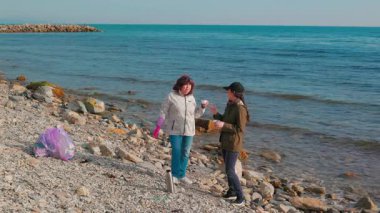 Gönüllüler, kirliliği önlemek ve sürdürülebilir yaşamı desteklemek amacıyla vahşi bir sahilden plastik atık ve diğer çöpleri topluyorlar. Kadınlar bir fren yapar ve çay içerler. Tam boy çekim.