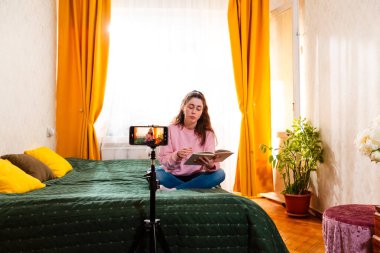 Kadın içerik yaratıcısı yatakta oturuyor sosyal medya vlog 'u için bir video kaydediyor tripoddaki akıllı telefonu kullanarak dijital markasını oluşturuyor. Geniş açı.