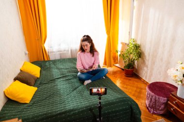 Beyaz kadın bir yatakta oturuyor ve akıllı telefon ve tripodu kullanarak bir vlog yaratıyor, sosyal medya ve online iş stratejilerine odaklanıyor. Geniş açı.