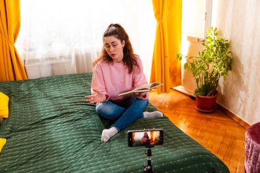 Konuşan bir kadın yatakta oturmuş, akıllı telefonuyla video kaydediyor, çevrimiçi takipçileri için yüksek kaliteli sosyal medya içeriği yaratıyor. Geniş açı.