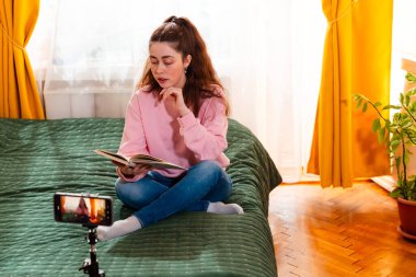 Tarz sahibi genç bir kadın kitap okuyor ve yatağında oturup sosyal medya takipçilerinin ilgisini çeken bir video çekiyor. Orta çekim.