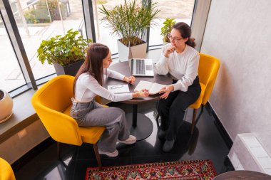 Akıllı kıyafetler giymiş iş kadınları arasında kahve içerken hedefe yönelik bir konuşma. Sakin, profesyonel ortam. Uzaktan işbirliği ve planlama başarısı kavramı. Üst manzara, geniş açı.