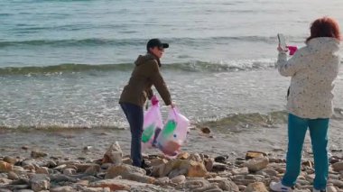 İki kadın eylemci, kirliliği önlemek ve doğayı korumak için plastik ve diğer döküntüleri toplayarak vahşi bir kıyı şeridini temizliyor. İnsanlar çevresel sorumluluk çabalarına katılıyorlar.