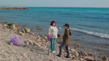 İki kadın aktivist bir plaj temizliğine katılarak kirliliği azaltmak için plastik atık topluyor. Gönüllüler bir fren yapıp çay içsin. Tam boy çekim
