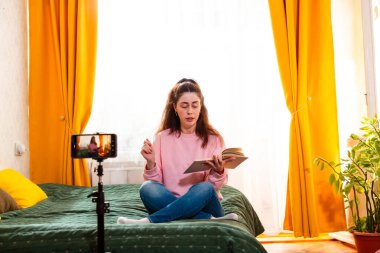 Kadın video kaydedicisi, akıllı telefon ve tripod kurulumu kullanarak bir yatakta otururken çevrimiçi kanalı için bir video kaydediyor. Geniş açı, düşük açı.