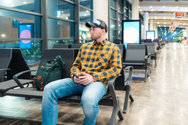Beyaz şapkalı ve ceketli yetişkin bir adamın havaalanındaki bekleme salonunda sandalyede otururken çekilmiş resmi. Uçuştan önce terminalde biniş kavramı.