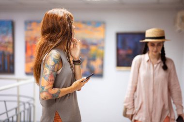 Kafkas tenli genç bir kadın. Dövmesi var. Tablolar üzerinde düşünüyor. Arka planda sanat galerisi sergi salonu var. Kültürel eğitim kavramı.
