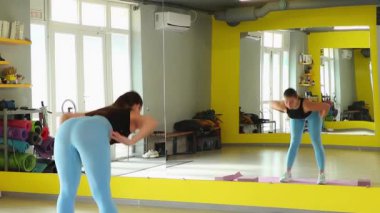 Genç, fit, beyaz bir kadın modern bir spor salonunda antrenman yapıyor. Aynanın yanında güç egzersizleri yapıyor. Kavram, sağlıklı bir yaşam tarzını ve fiziksel disiplini yansıtır. Arka plan, geniş açı.