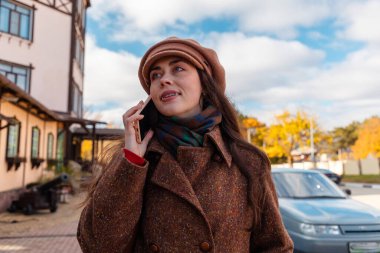 Sonbahar günü şehir caddesinde, paltolu ve şapkalı gülümseyen güzel beyaz bir kadının alt manzarası. Yaşam tarzı ve şehir hayatı.