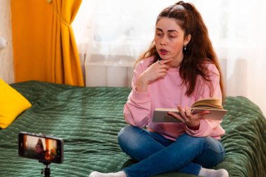 Sayısal içerik yaratıcısı rahat bir yatakta oturup üç ayaklı bir telefondan akıllı bir video bloğu çekiyor ve çevrimiçi eğilimleri tartışıyor. Kadın kitap okuyor..