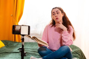 Çevrimiçi koç yatakta oturuyor ve sosyal medya için motive edici bir video bloğu çekiyor, tripodun üzerinde akıllı telefon kullanıyor. Orta çekim.