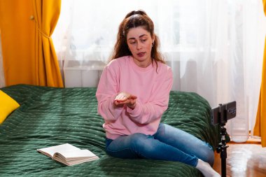 Çevrimiçi içerik yaratıcısı rahat bir yatakta oturuyor ve bir video bloğu çekiyor, izleyicisiyle iletişim kurmak için akıllı telefon kullanıyor. Orta çekim.