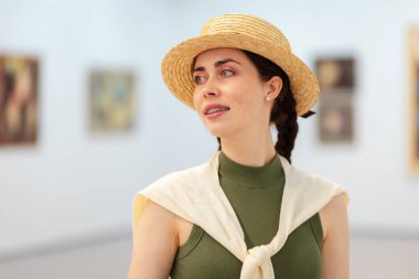 Sanat galerisinde poz veren saman şapkalı, genç, beyaz, zarif bir kadının portresi. Resimler arka planda. Kültür Eğitimi ve Müze Günü kavramı.