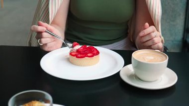 Güneşli bir kafe köşesinde oturan bir kadın sıcak kapuçinosunun ve zengin katmanlı kekinin tadını çıkarıyor. Kadınlığın, lezzetin ve şatafatlı barışın sahnesi