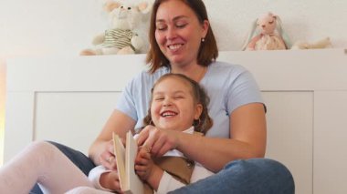 Genç beyaz kadın, rahat bir yatak odasında kitap okurken küçük kızına sarılıyor. Evde erken eğitim ve çocuk gelişimi kavramı