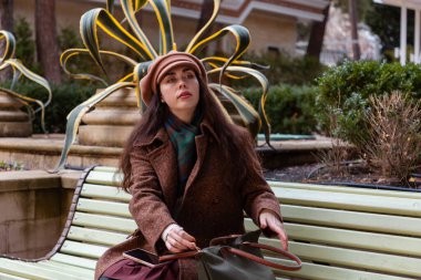 Kürklü zarif bir kadın çantasıyla bankta oturuyor ve şehirde sakin bir anın tadını çıkarıyor. Kadın şehir yaşam tarzı, rahatlama, turizm ve şehir atmosferi