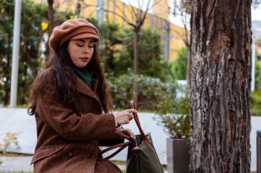 Sakin bir şehir yürüyüşü sırasında parktaki bankta çantasıyla dinlenen şık beyaz bir kadın. Rahatlama, sokak modası ve kentsel çevrede günlük kadın yaşam tarzı