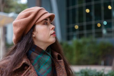 Açık havada cam bir binanın yanında duran şapkalı ve eşarplı beyaz bir kadının portresi. Şehir yürüyüşleri