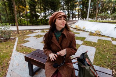 Paltolu genç beyaz kadın kentteki bir bankta tek başına oturuyor, yanında çantasıyla rahatlıyor. Şehir hayatı, hafta sonu tatili ve huzurlu bir açık hava anı.