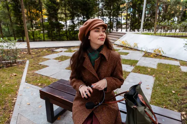 Paltolu genç beyaz kadın kentteki bir bankta tek başına oturuyor, yanında çantasıyla rahatlıyor. Şehir hayatı, hafta sonu tatili ve huzurlu bir açık hava anı.