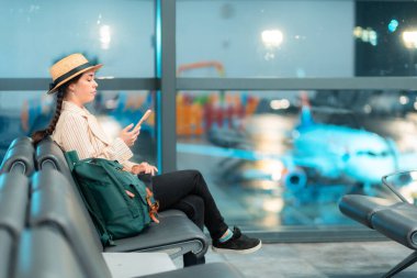 Şapka ve ceket giyen beyaz bir kadının havaalanındaki bekleme salonunda sandalyede otururken çekilmiş geniş bir fotoğrafı. Fotokopi uzayı ile seyahat afişi. Uçuştan önce terminalde biniş kavramı.