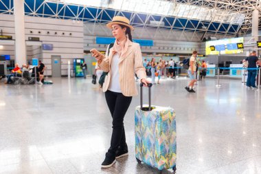 Zarif takım elbiseli beyaz bir kadının geniş açılı resmi akıllı telefon kullanıyor ve bavul tutuyor. Havaalanında biri var. Seyahat ve seyahat kavramı