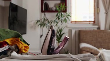 Buharlı kızgın demir, ütü masasının üstünde duruyor, yakın plan. Arka planda rahat bir iç mekan. Ev işleri ve çamaşır..