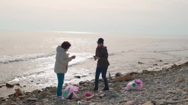 İki çevreci kirlenmiş bir plajdan atık topluyor. Gönüllüler doğayı temizlemek ve bilinçlendirmek için birlikte çalışırlar. Kadınlar fotoğraf çeker. Geniş açı.